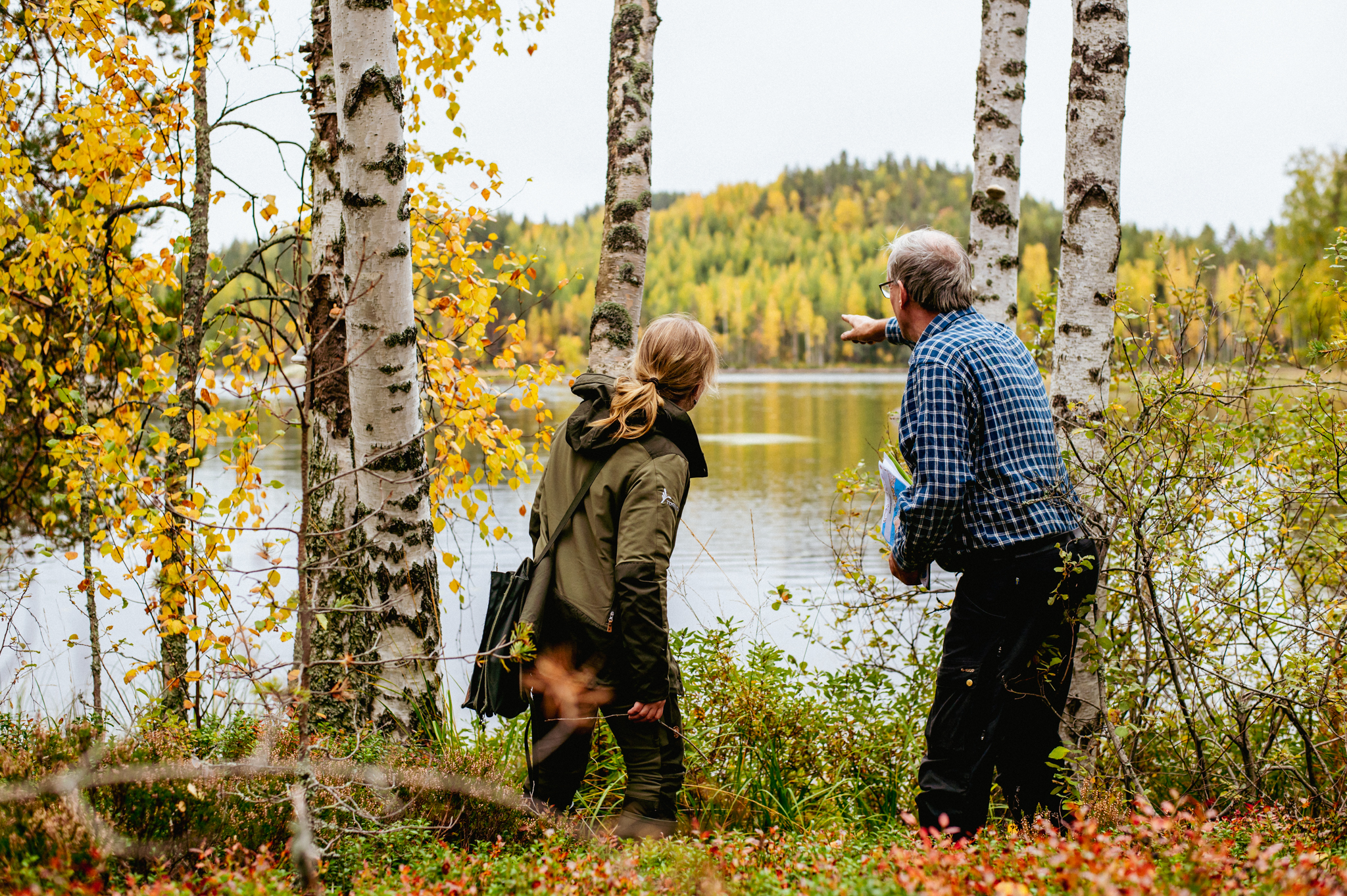 Mies ja nainen metsän reunalla järven rannalla, mies osoittaa vastarannan metsää.